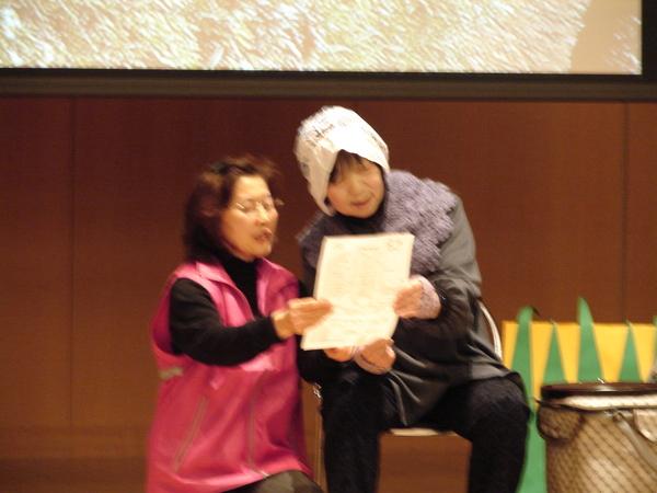 椅子に腰かけているおばあさんと一緒に書類を手に持ちお話ししている写真